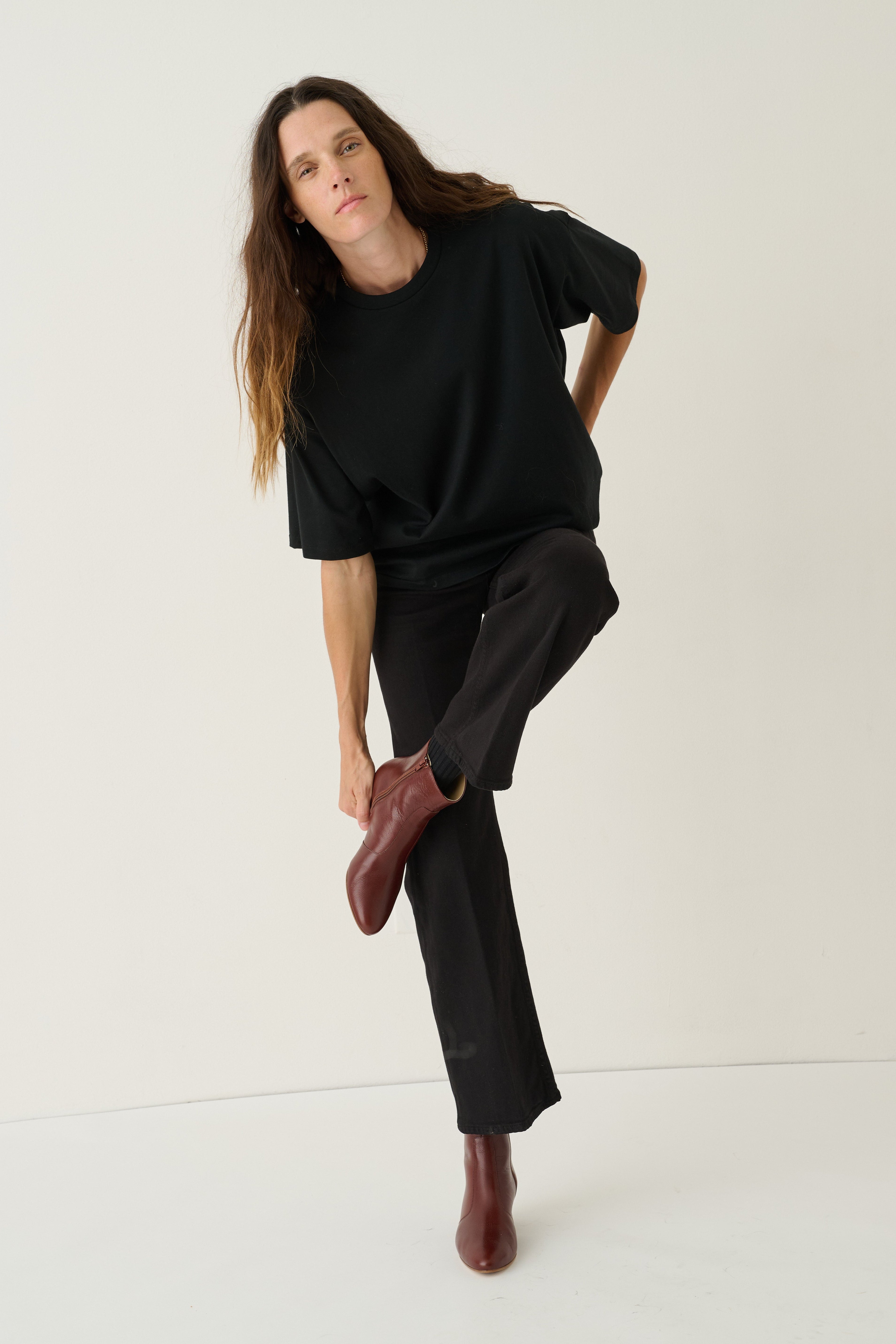 A woman modeling a black oversized short-sleeve top and black wide-leg trousers, paired with dark brown leather ankle boots, against a plain off-white studio background.