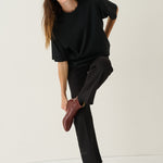 A woman modeling a black oversized short-sleeve top and black wide-leg trousers, paired with dark brown leather ankle boots, against a plain off-white studio background.