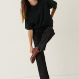 A woman modeling a black oversized short-sleeve top and black wide-leg trousers, paired with dark brown leather ankle boots, against a plain off-white studio background.
