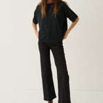 A woman modeling a black short-sleeve t-shirt and black wide-leg cropped pants with brown ankle boots, standing against a plain off-white studio background.