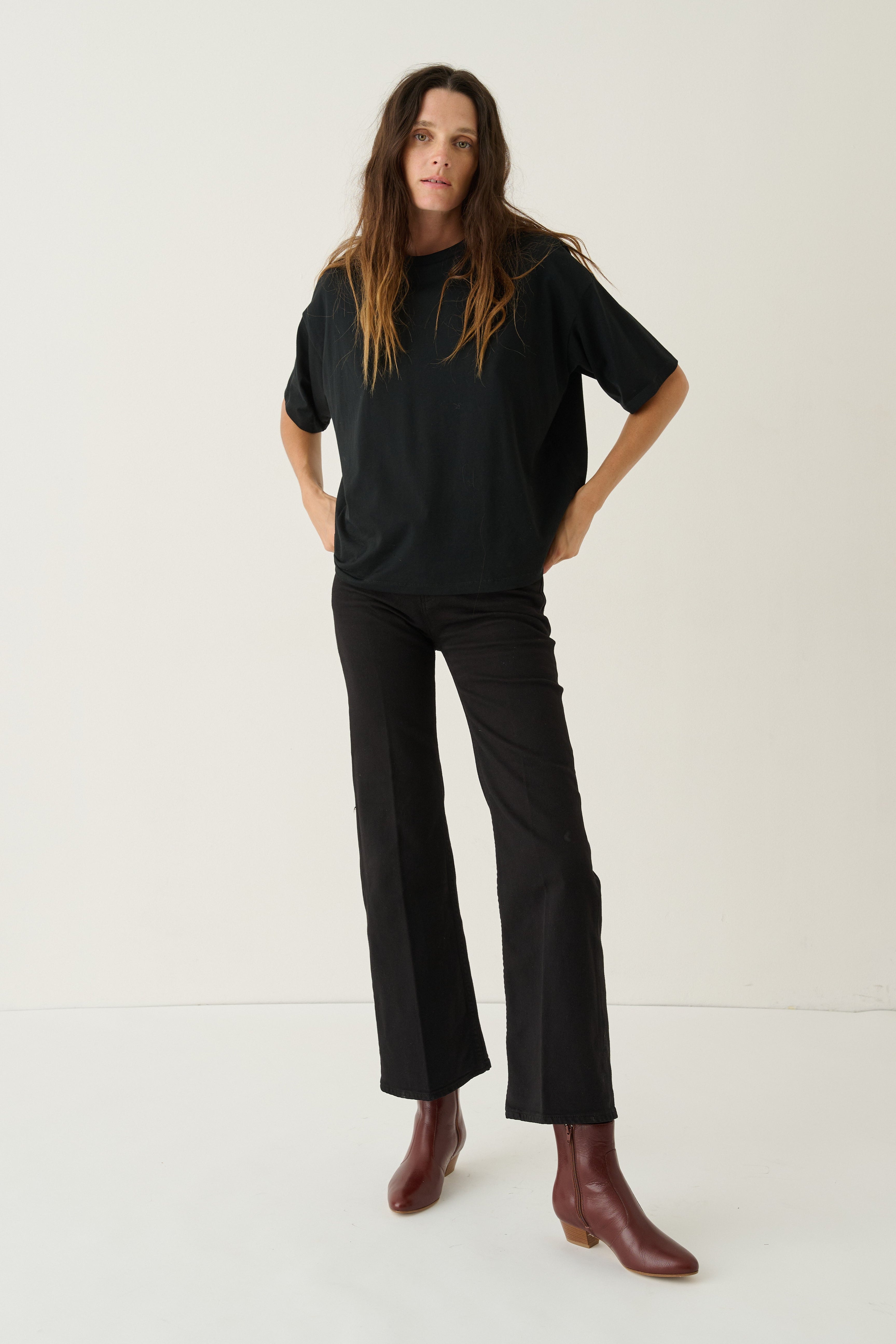 A woman modeling a black short-sleeve t-shirt and black wide-leg cropped pants with brown ankle boots, standing against a plain off-white studio background.