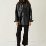 A model stands against a plain off-white studio background wearing a black oversized faux-leather button-up shirt jacket, black trousers, and brown ankle boots.