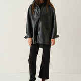 A model stands against a plain off-white studio background wearing a black oversized faux-leather button-up shirt jacket, black trousers, and brown ankle boots.