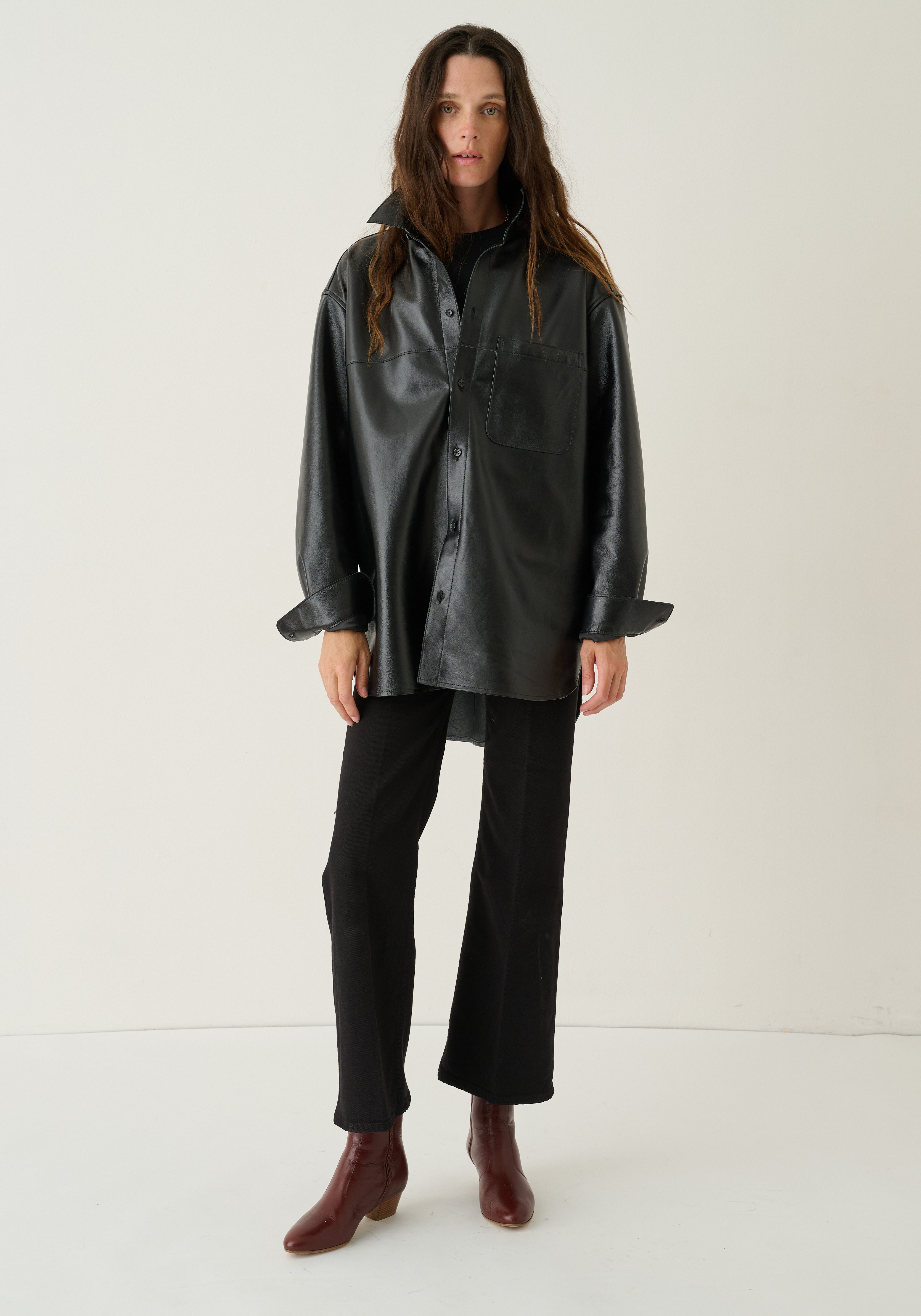 A model stands against a plain off-white studio background wearing a black oversized faux-leather button-up shirt jacket, black trousers, and brown ankle boots.