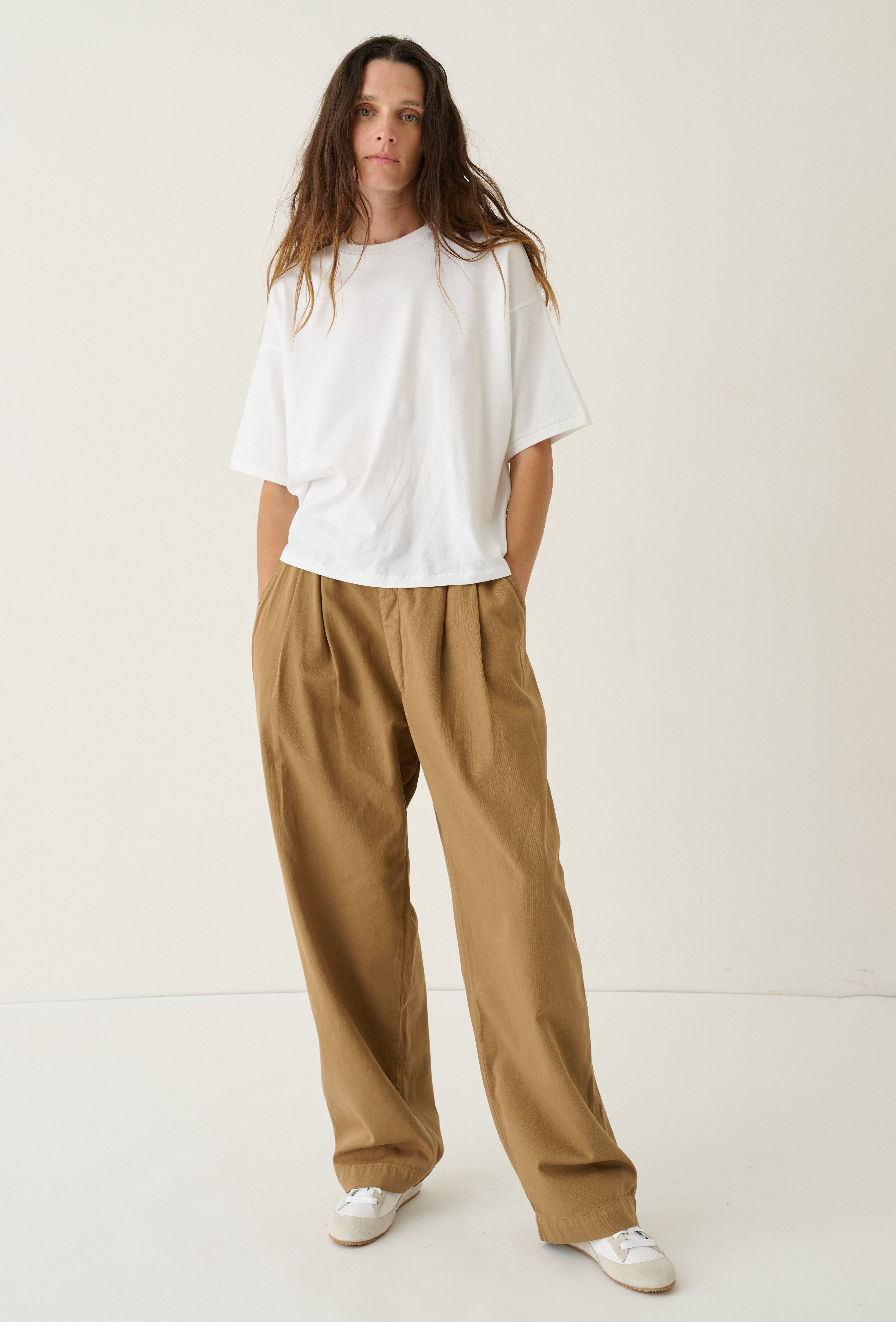 A woman modeling a white short-sleeve t-shirt and loose, wide-leg brown pants against a plain, light beige studio background.