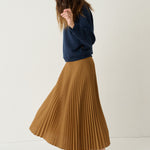 A woman wearing a navy blue sweatshirt, a pleated mustard-brown midi skirt, and burgundy loafers, standing against a plain off-white studio background.