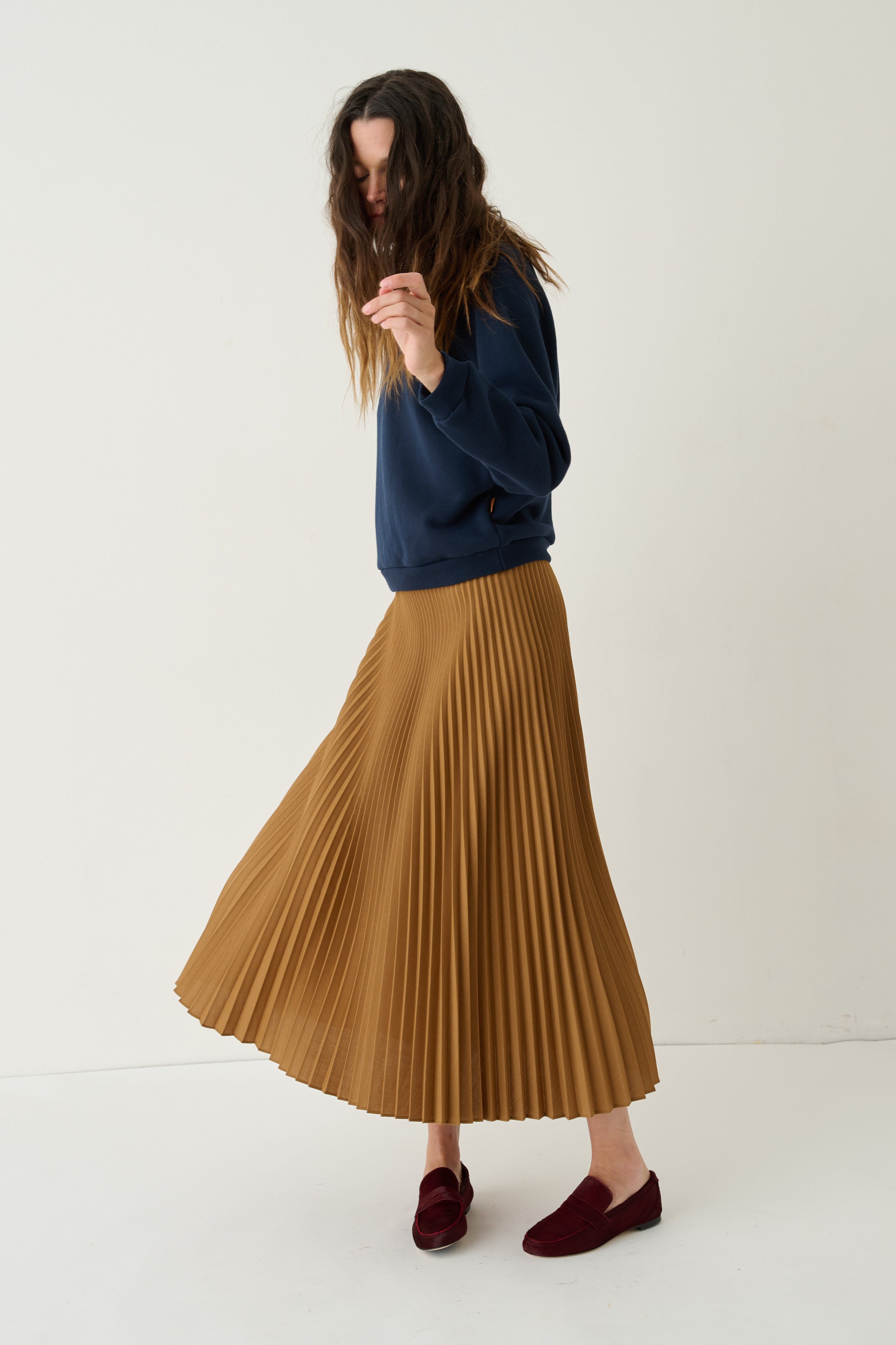 A woman wearing a navy blue sweatshirt, a pleated mustard-brown midi skirt, and burgundy loafers, standing against a plain off-white studio background.