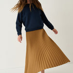 A woman wearing a navy blue sweatshirt, a pleated mustard yellow midi skirt, and burgundy loafers, captured in motion against a clean off-white studio background.