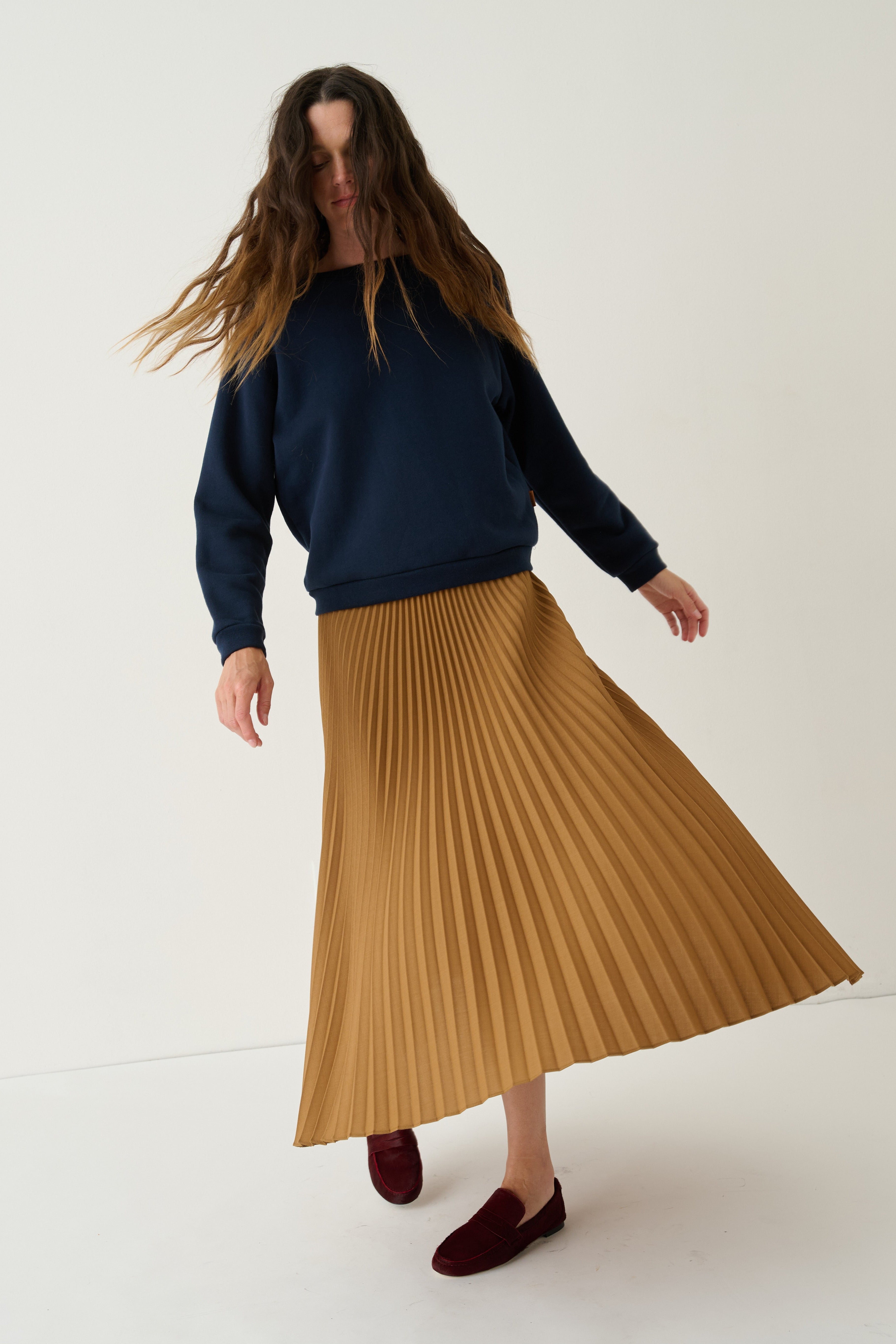A woman wearing a navy blue sweatshirt, a pleated mustard yellow midi skirt, and burgundy loafers, captured in motion against a clean off-white studio background.