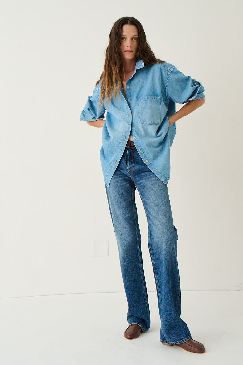 A woman modeling an oversized light blue denim button-up shirt and matching blue jeans with brown shoes, standing against a plain off-white studio background.