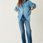 A woman modeling an oversized light blue denim button-up shirt and matching blue jeans with brown shoes, standing against a plain off-white studio background.