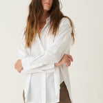 A woman modeling a white long-sleeve button-up shirt and brown cropped trousers with burgundy heels, standing against a plain off-white studio background.