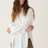 A woman modeling a white long-sleeve button-up shirt and brown cropped trousers with burgundy heels, standing against a plain off-white studio background.