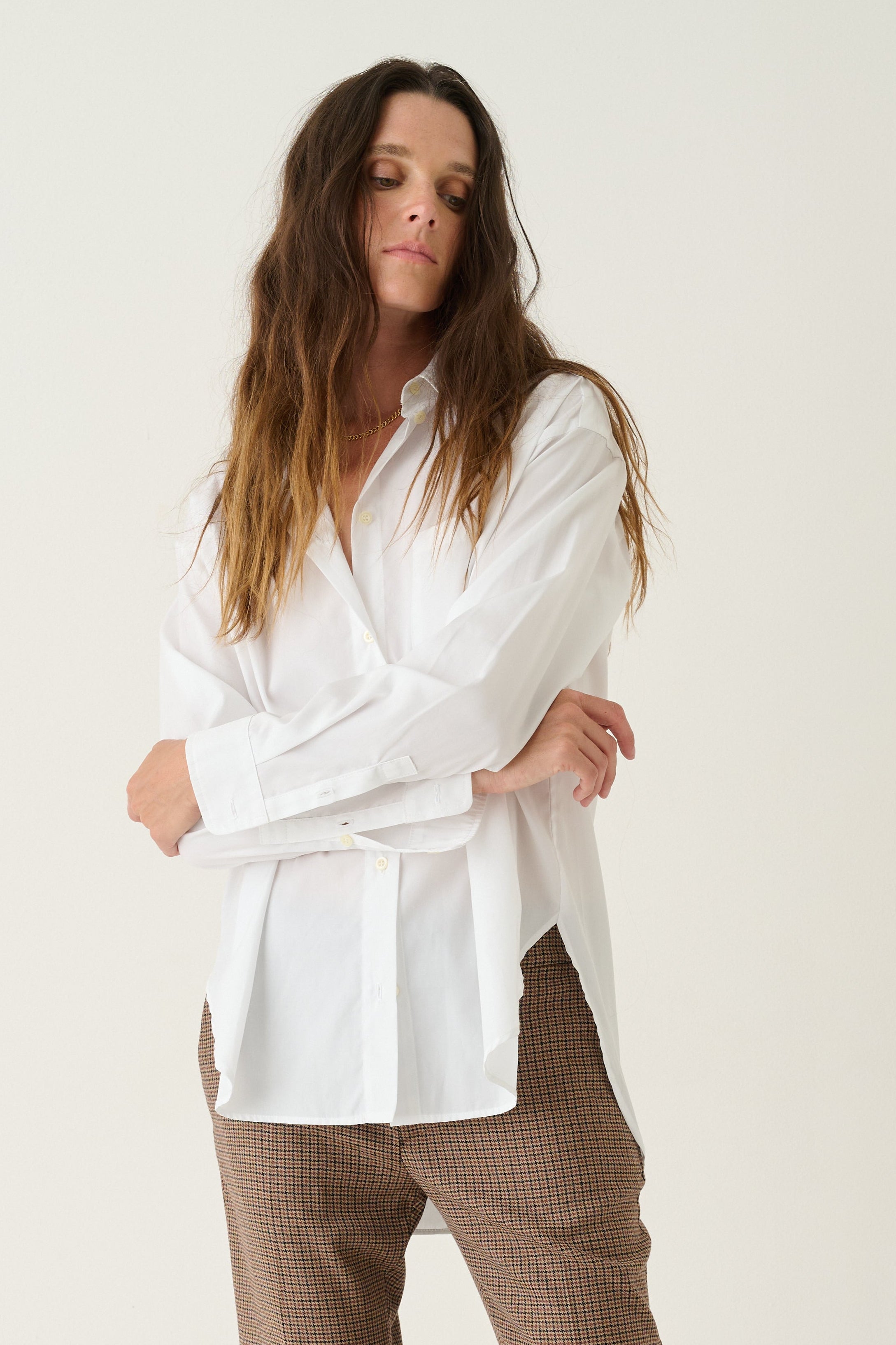 A woman modeling a white long-sleeve button-up shirt and brown cropped trousers with burgundy heels, standing against a plain off-white studio background.
