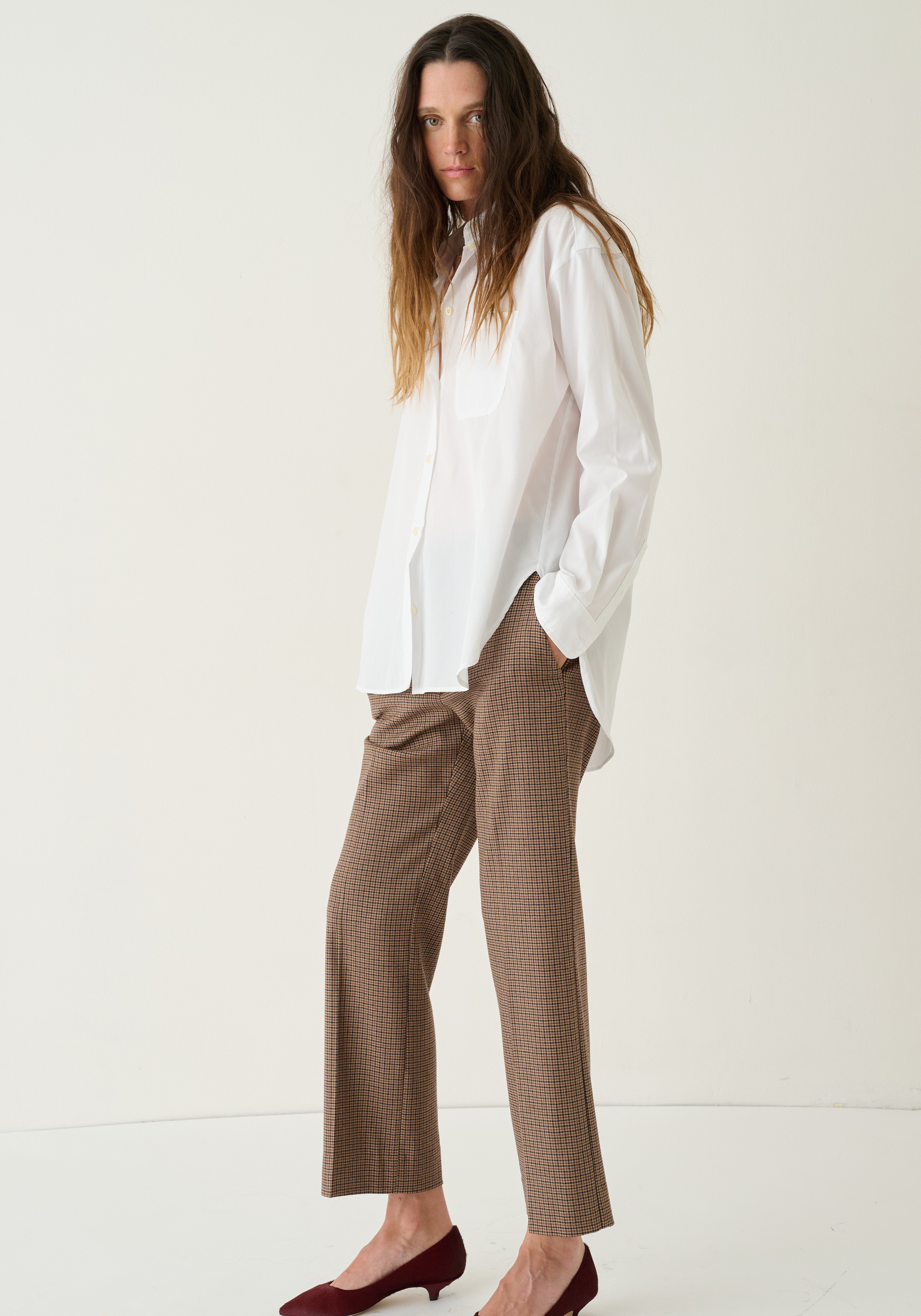 A woman modeling a white long-sleeve button-up shirt and brown houndstooth cropped trousers with burgundy pointed-toe flats, set against a plain off-white studio background.