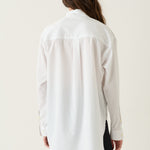 A woman viewed from behind wearing a loose white long-sleeve button-up shirt and black pants, standing against a plain off-white studio background.