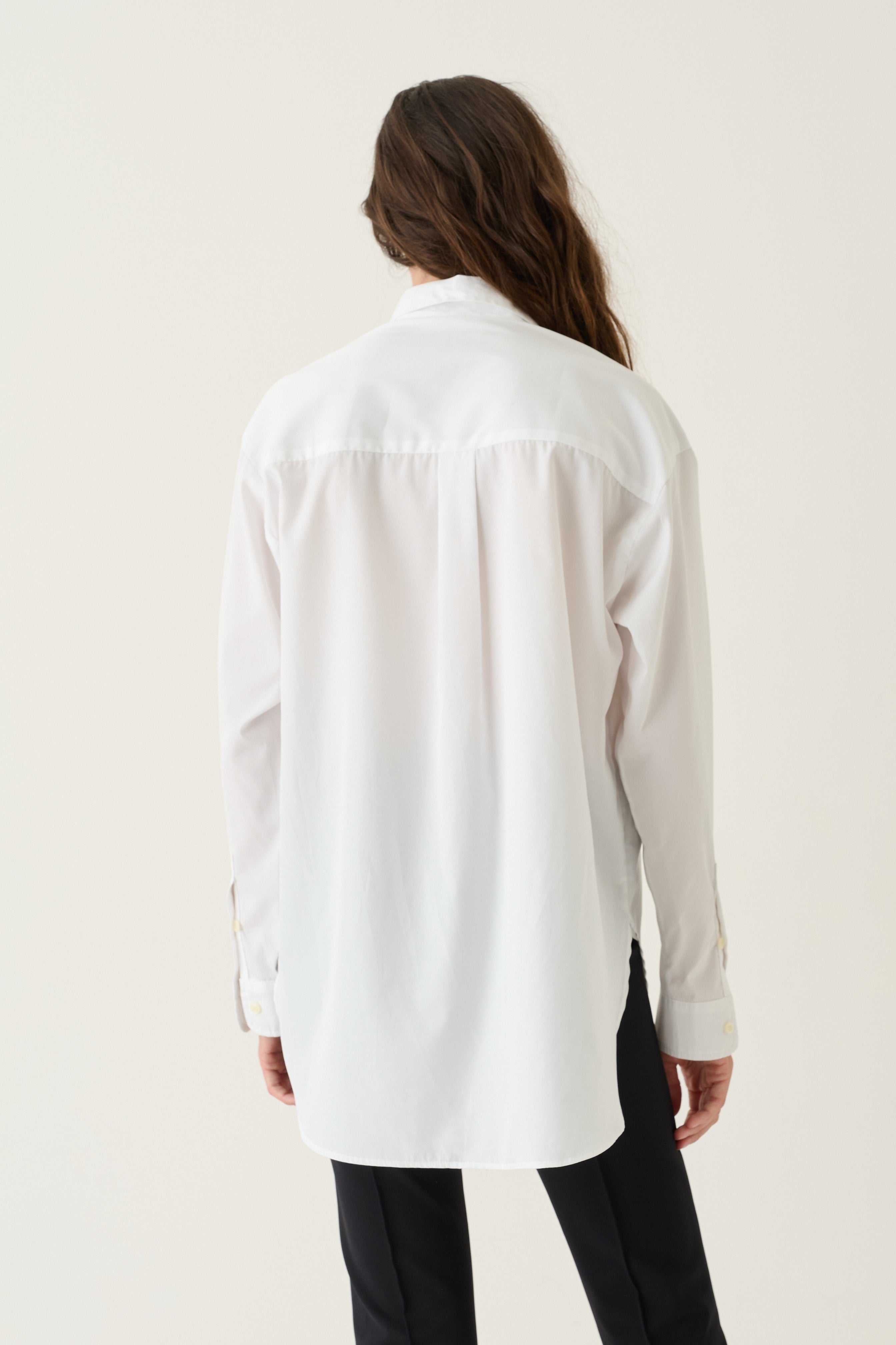 A woman viewed from behind wearing a loose white long-sleeve button-up shirt and black pants, standing against a plain off-white studio background.