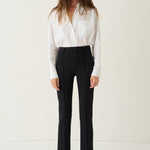A woman stands against a plain off-white studio background wearing a white long-sleeve button-up shirt, black high-waisted cropped trousers, and black shoes.