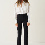 A woman stands against a plain off-white studio background wearing a white long-sleeve button-up shirt, black high-waisted cropped trousers, and black shoes.
