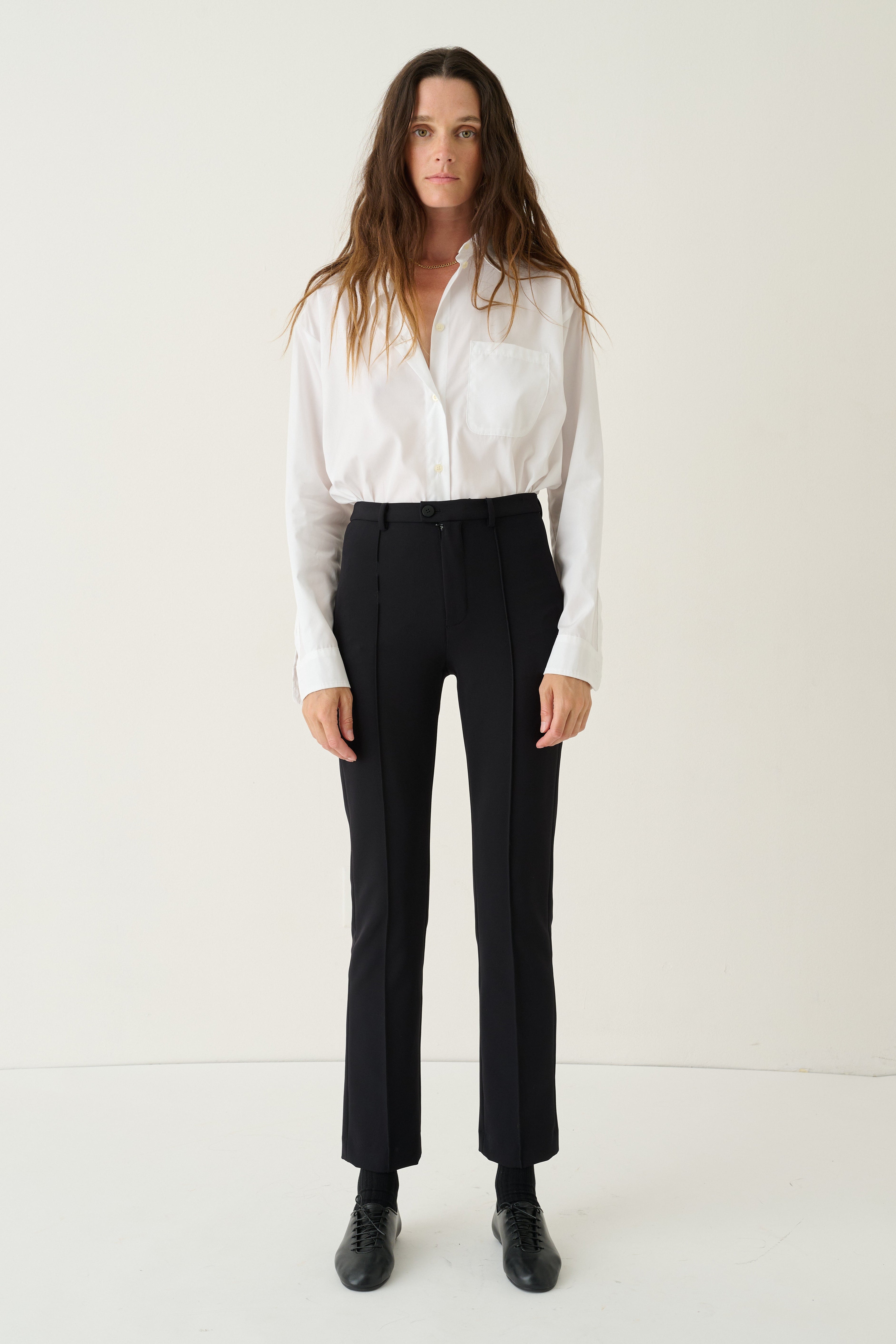 A woman stands against a plain off-white studio background wearing a white long-sleeve button-up shirt, black high-waisted cropped trousers, and black shoes.