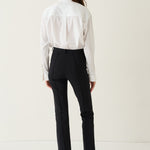 A model shown from the back wearing a white long-sleeve blouse and black trousers, standing against a plain off-white studio background.