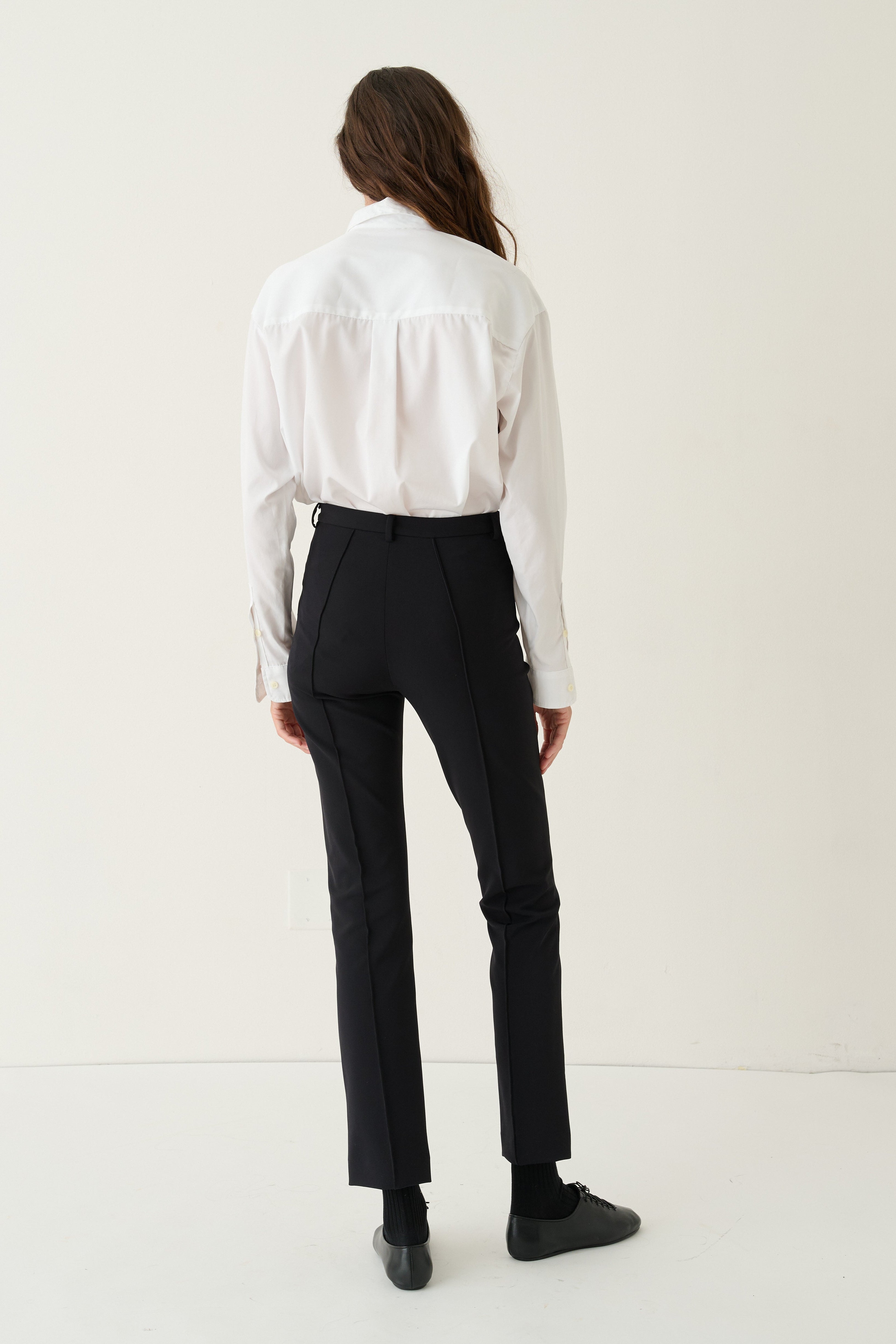 A model shown from the back wearing a white long-sleeve blouse and black trousers, standing against a plain off-white studio background.