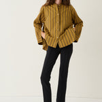 A woman modeling a mustard yellow button-up shirt with thin black vertical stripes, paired with black trousers and black shoes, standing against a plain white studio background.