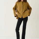 A woman modeling a mustard yellow button-up shirt with thin black vertical stripes, paired with black trousers and black shoes, standing against a plain white studio background.
