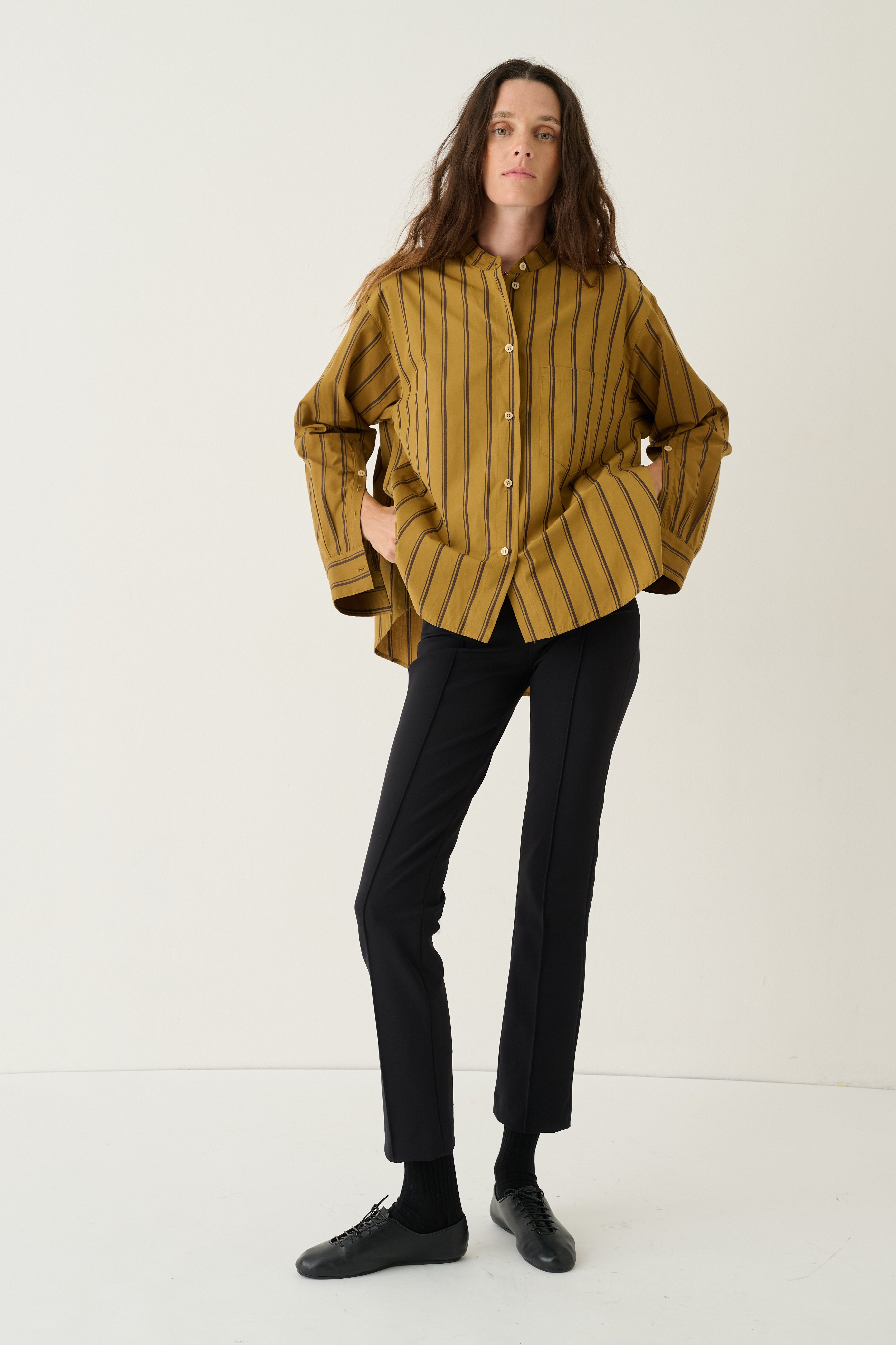 A woman modeling a mustard yellow button-up shirt with thin black vertical stripes, paired with black trousers and black shoes, standing against a plain white studio background.