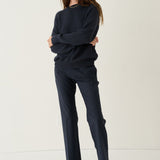 A woman modeling a dark navy knit sweater and matching navy trousers with brown leather shoes, standing against a plain off-white studio background.