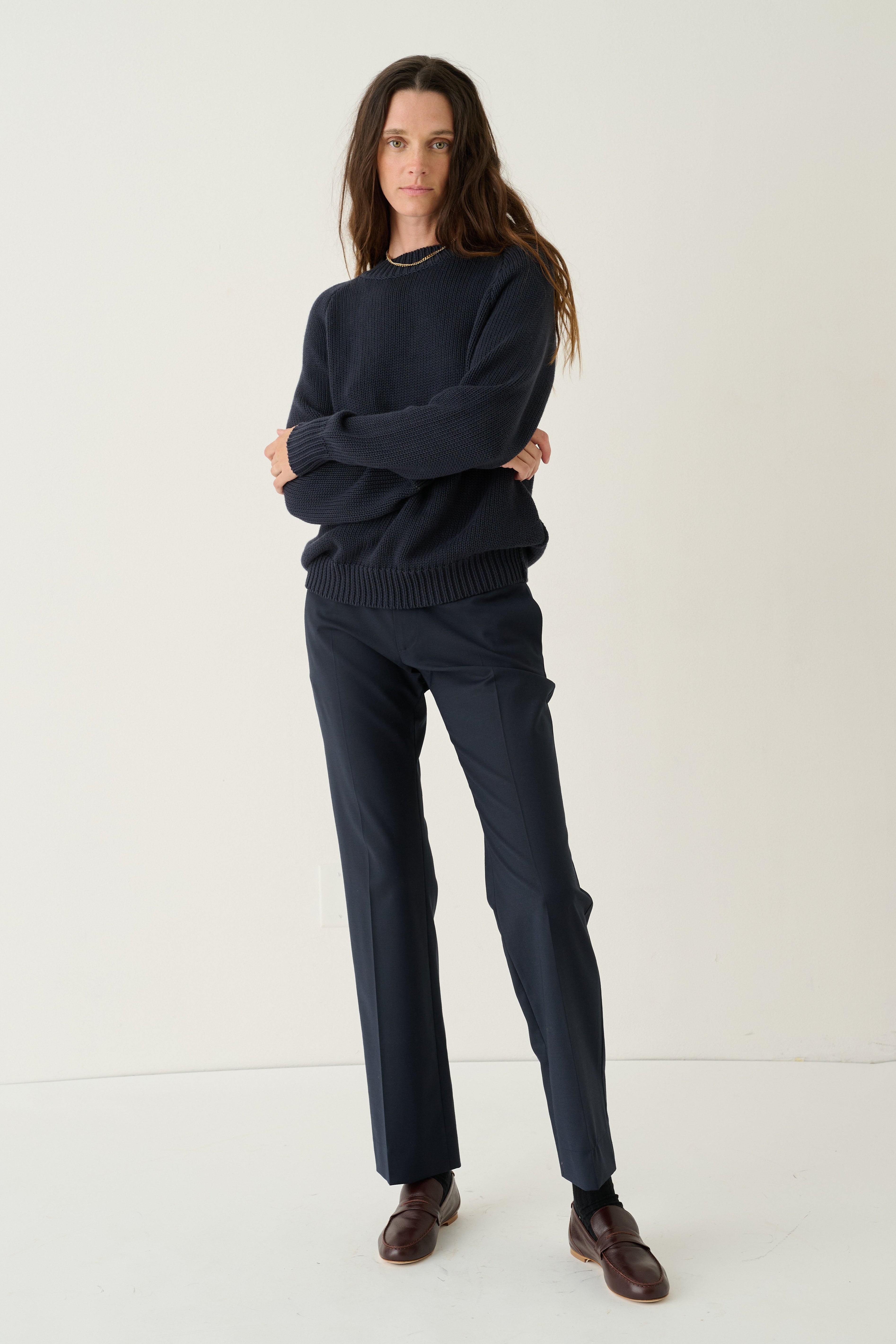 A woman modeling a dark navy knit sweater and matching navy trousers with brown leather shoes, standing against a plain off-white studio background.