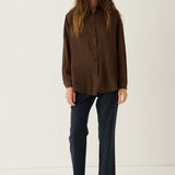 A woman stands against a plain off-white studio background wearing a dark brown long-sleeve button-up shirt, dark navy trousers, and dark brown leather loafers.