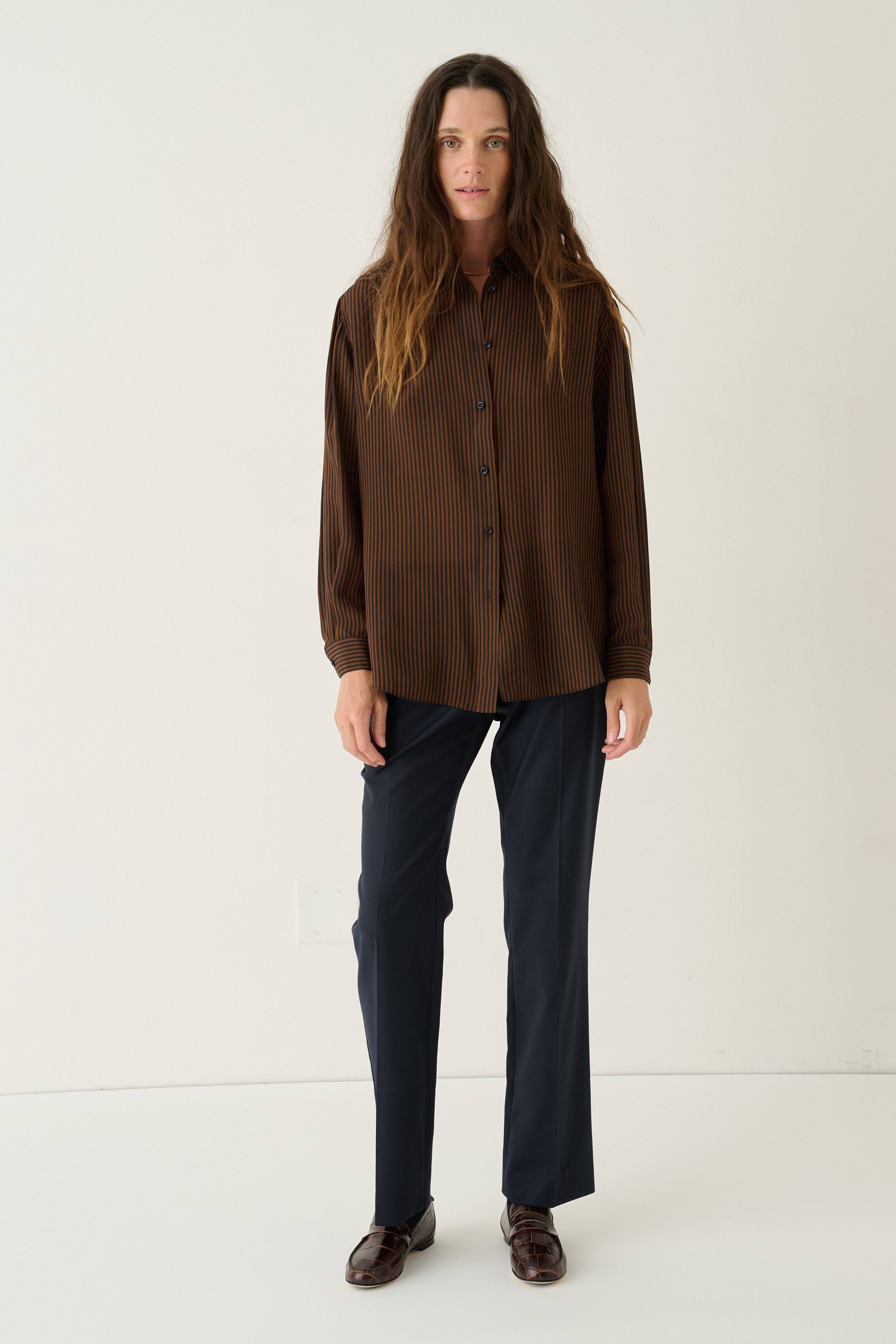 A woman stands against a plain off-white studio background wearing a dark brown long-sleeve button-up shirt, dark navy trousers, and dark brown leather loafers.