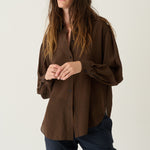 A woman modeling a brown pleated button-up shirt with dark navy pants, standing against a plain light gray studio background.