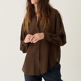 A woman modeling a brown pleated button-up shirt with dark navy pants, standing against a plain light gray studio background.