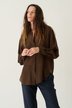 A woman modeling a brown pleated button-up shirt with dark navy pants, standing against a plain light gray studio background.