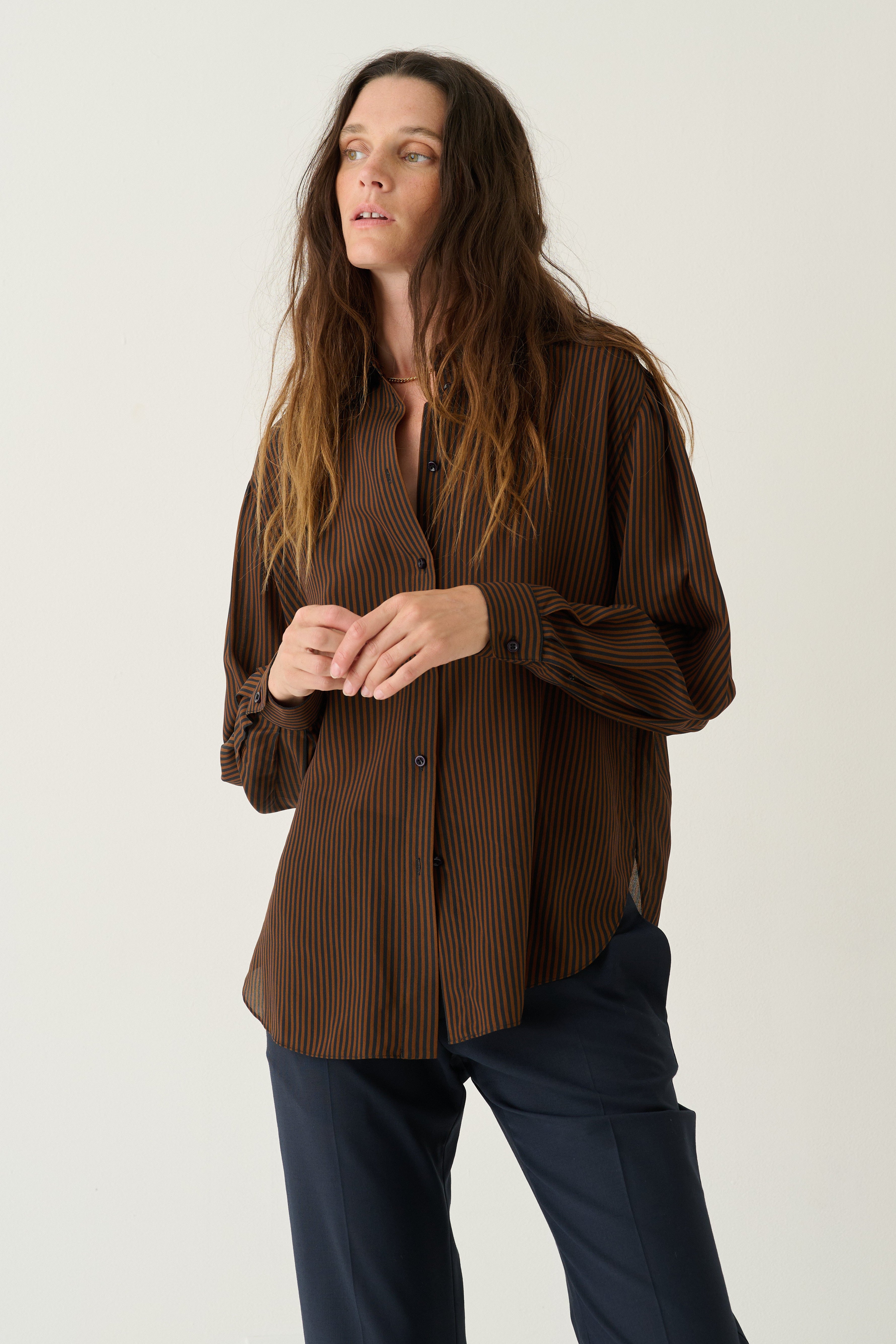 A woman modeling a brown pleated button-up shirt with dark navy pants, standing against a plain light gray studio background.