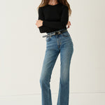 A woman modeling a pair of high-waisted, straight-leg blue jeans with a black belt, paired with a black long-sleeve top and black leather shoes, against a plain white studio background.