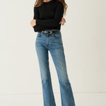 A woman modeling a pair of medium-wash blue flare jeans with a black belt, paired with a black long-sleeve top and black leather shoes, standing against a plain off-white studio background.