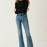 A woman modeling a pair of medium-wash blue flare jeans with a black belt, paired with a black long-sleeve top and black leather shoes, standing against a plain off-white studio background.