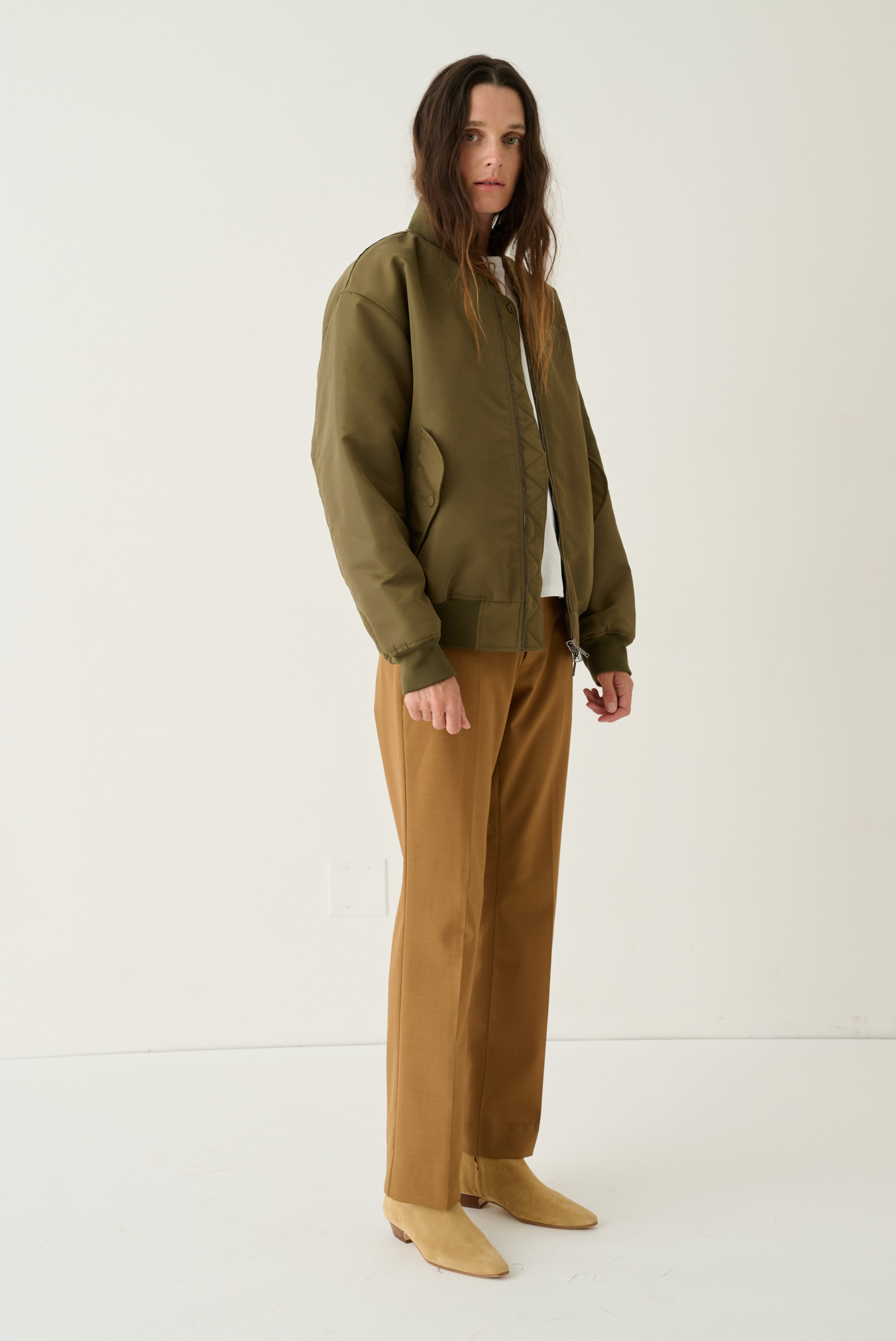 A model stands in a studio wearing an olive green bomber jacket over a white top, paired with brown wide-leg pants and tan pointed-toe flats.
