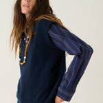 A woman modeling a navy blue ribbed knit sweater vest with long blue striped sleeves, paired with black flared pants and tan suede ankle boots, against a plain white studio background.