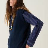 A woman modeling a navy blue ribbed knit sweater vest with long blue striped sleeves, paired with black flared pants and tan suede ankle boots, against a plain white studio background.