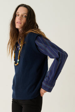 A woman modeling a navy blue ribbed knit sweater vest with long blue striped sleeves, paired with black flared pants and tan suede ankle boots, against a plain white studio background.
