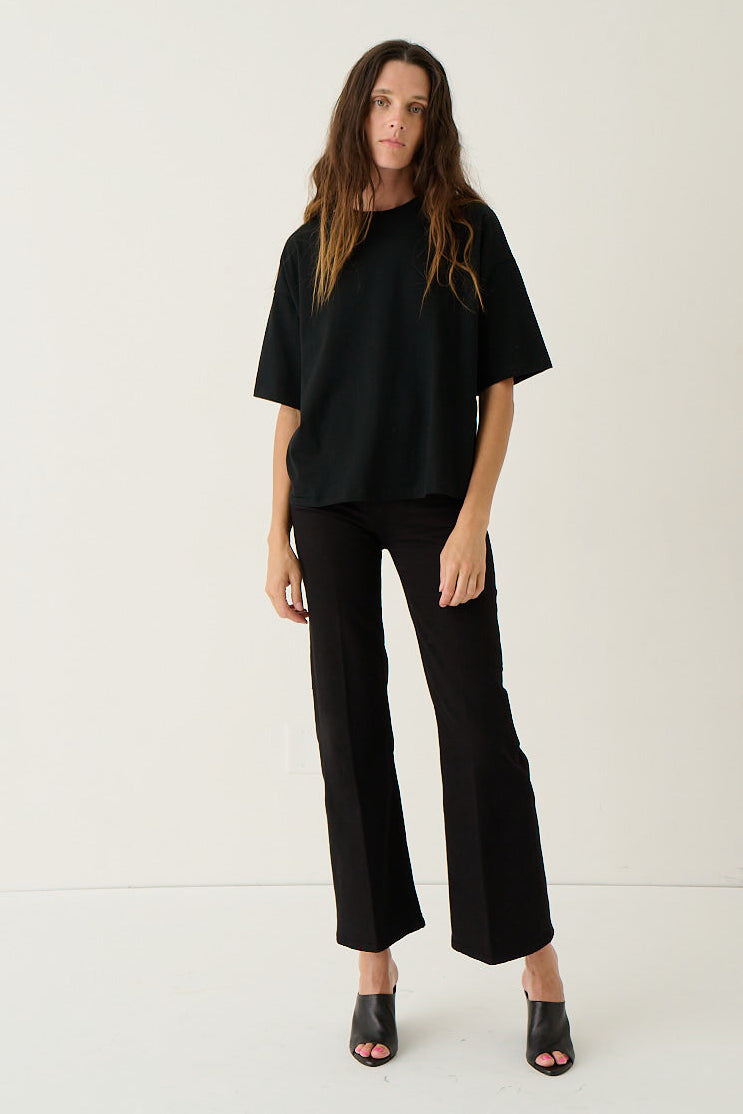 A woman modeling a black oversized short-sleeve top and black wide-leg cropped pants with black open-toe heels, standing against a plain off-white studio background.