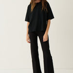 A woman modeling a black oversized short-sleeve top and black wide-leg cropped pants with black open-toe heels, standing against a plain off-white studio background.