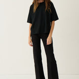 A woman modeling a black oversized short-sleeve top and black wide-leg cropped pants with black open-toe heels, standing against a plain off-white studio background.