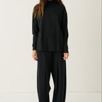 A woman modeling a black long-sleeve mock neck top and matching black wide-leg trousers, paired with white sneakers, against a plain off-white studio background.