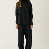 A woman modeling a black long-sleeve mock neck top and matching black wide-leg trousers, paired with white sneakers, against a plain off-white studio background.