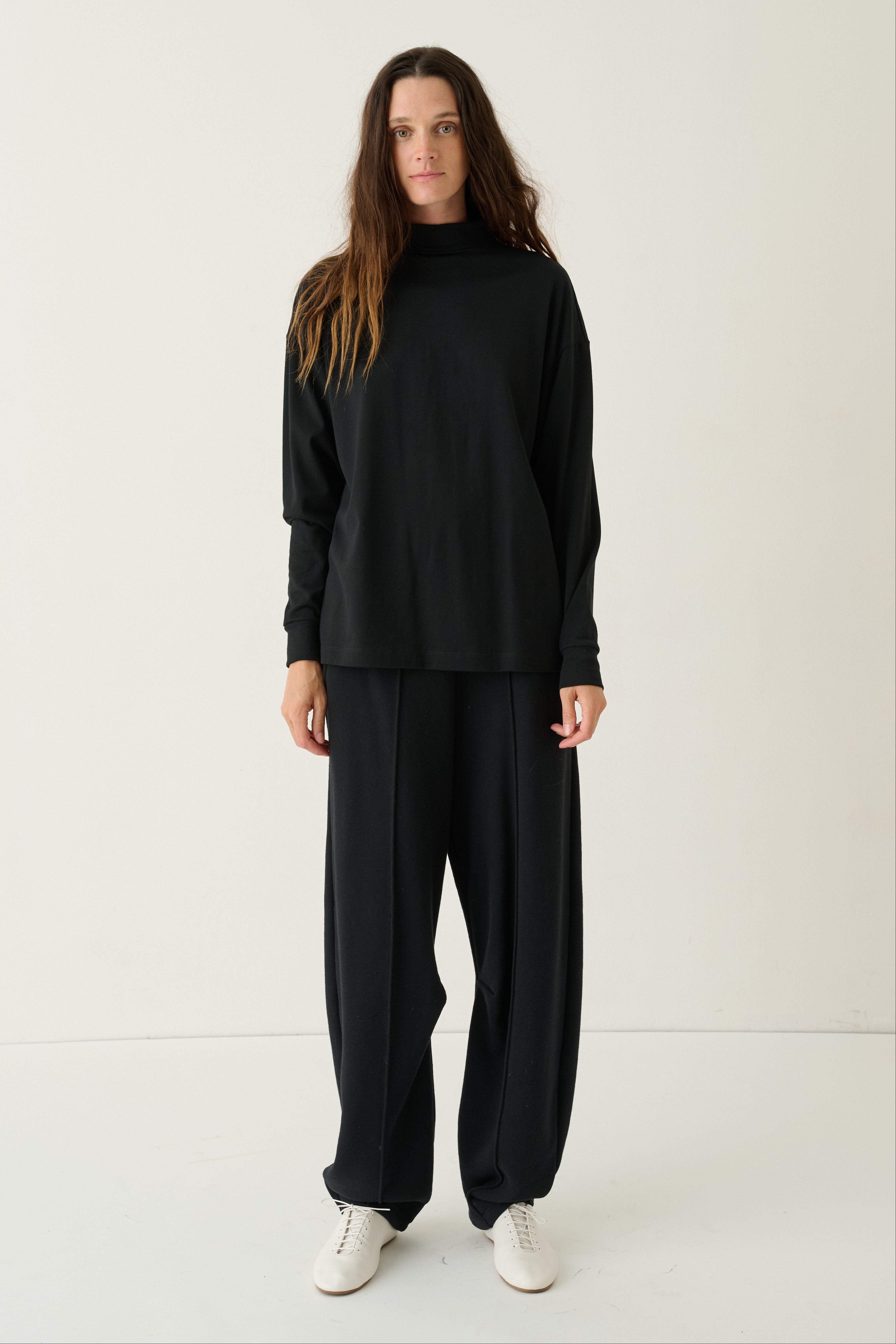 A woman modeling a black long-sleeve mock neck top and matching black wide-leg trousers, paired with white sneakers, against a plain off-white studio background.